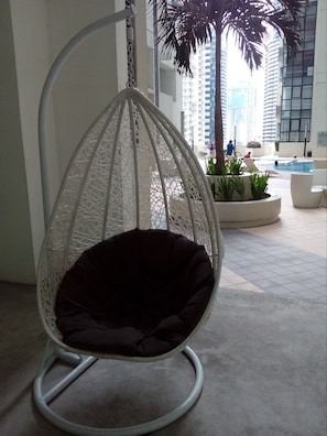 Una silla colgante con un cojín oscuro en un ambiente interior con piscina y edificios altos al fondo.