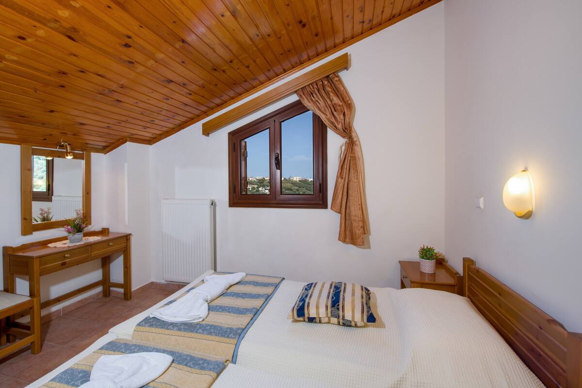 A bedroom with a wooden ceiling, a large window with curtains, a bedside table with a plant, a bed with a striped blanket, and a wooden desk with a mirror.