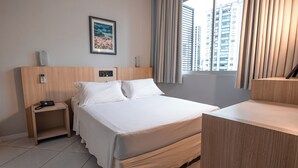 A hotel room with a large bed, a bedside table with a phone, a small shelf, a framed picture on the wall, and a window with curtains. - Sagres Praia Hotel (Balneário Camboriú)