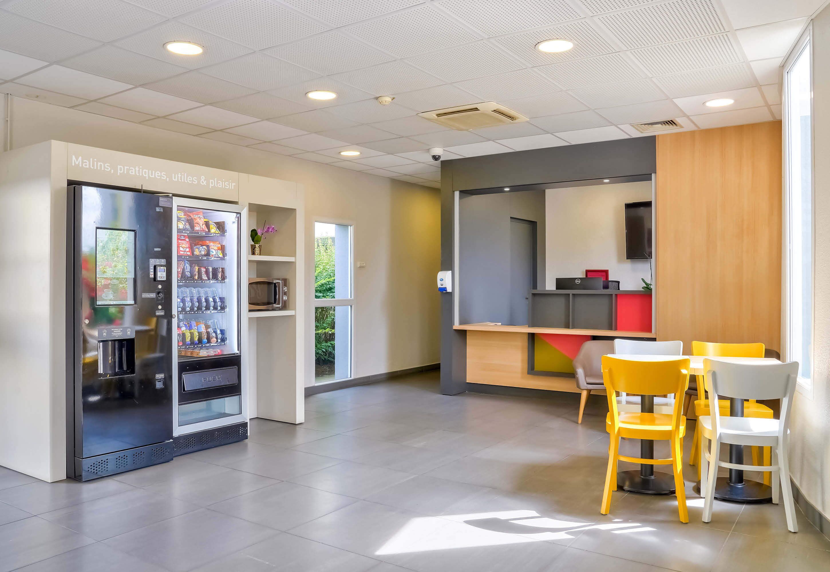 A modern indoor space with vending machines, a seating area with yellow and white chairs, and a counter with a red and gray color scheme.