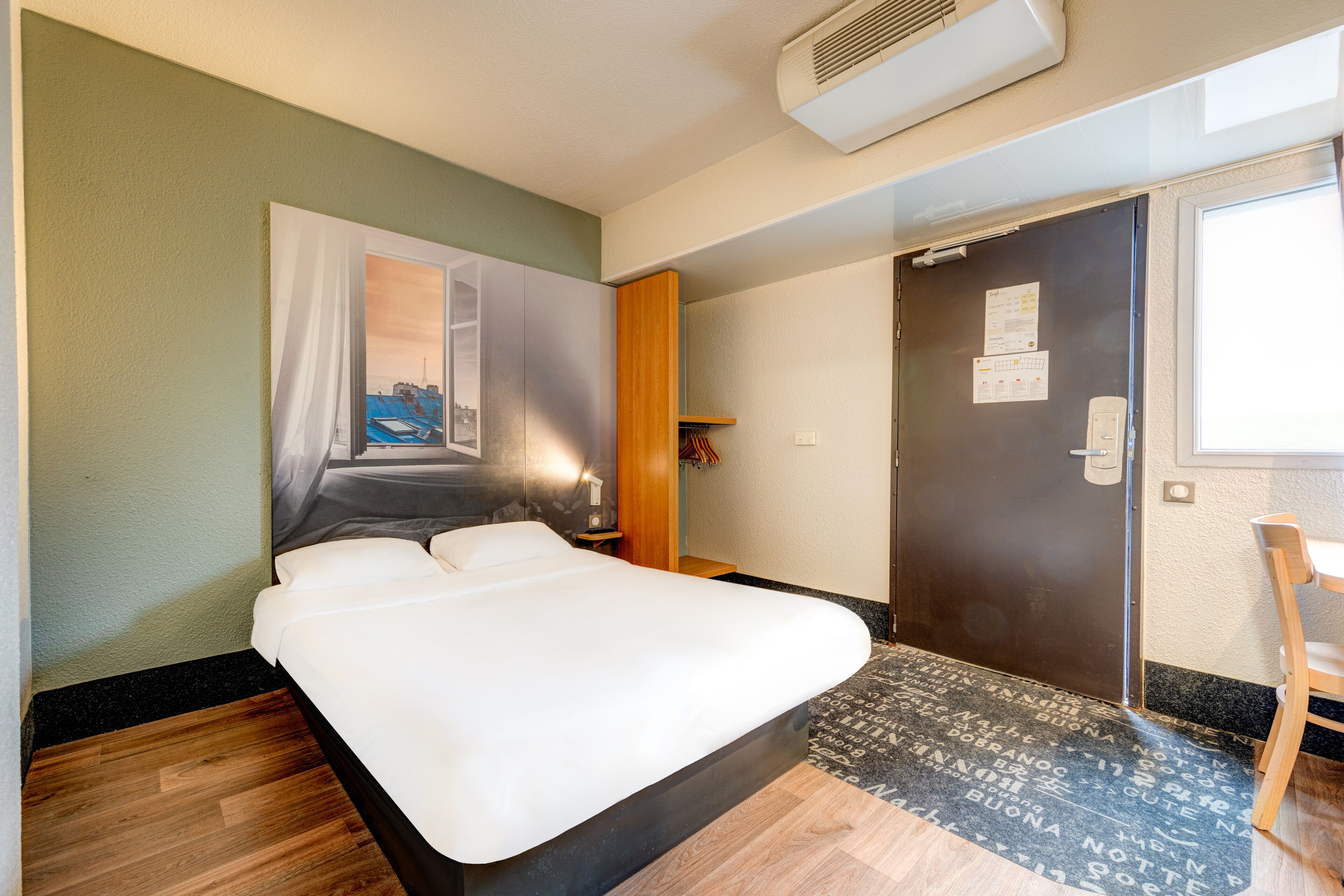 A hotel room with a bed, a wooden floor, a door, and a window.