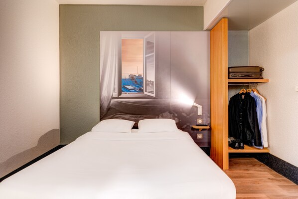Una cama bien hecha en una habitación con un ventanal grande, un armario de madera y un estante pequeño con una maleta y ropa.