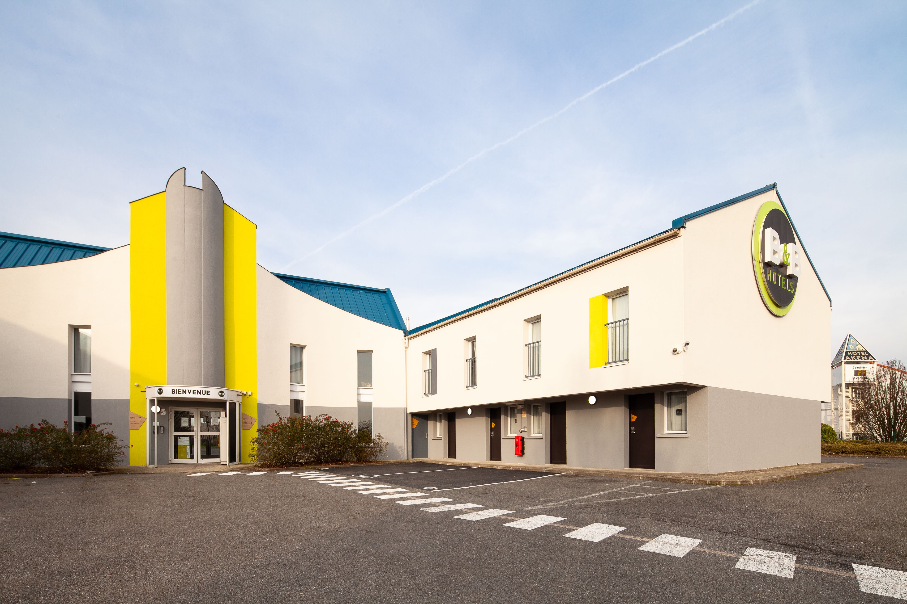 A modern hotel with a yellow and white facade, a 'Bienvenue' entrance, and a parking lot.