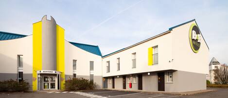A modern hotel with a yellow and white facade, a 'Bienvenue' entrance, and a parking lot.