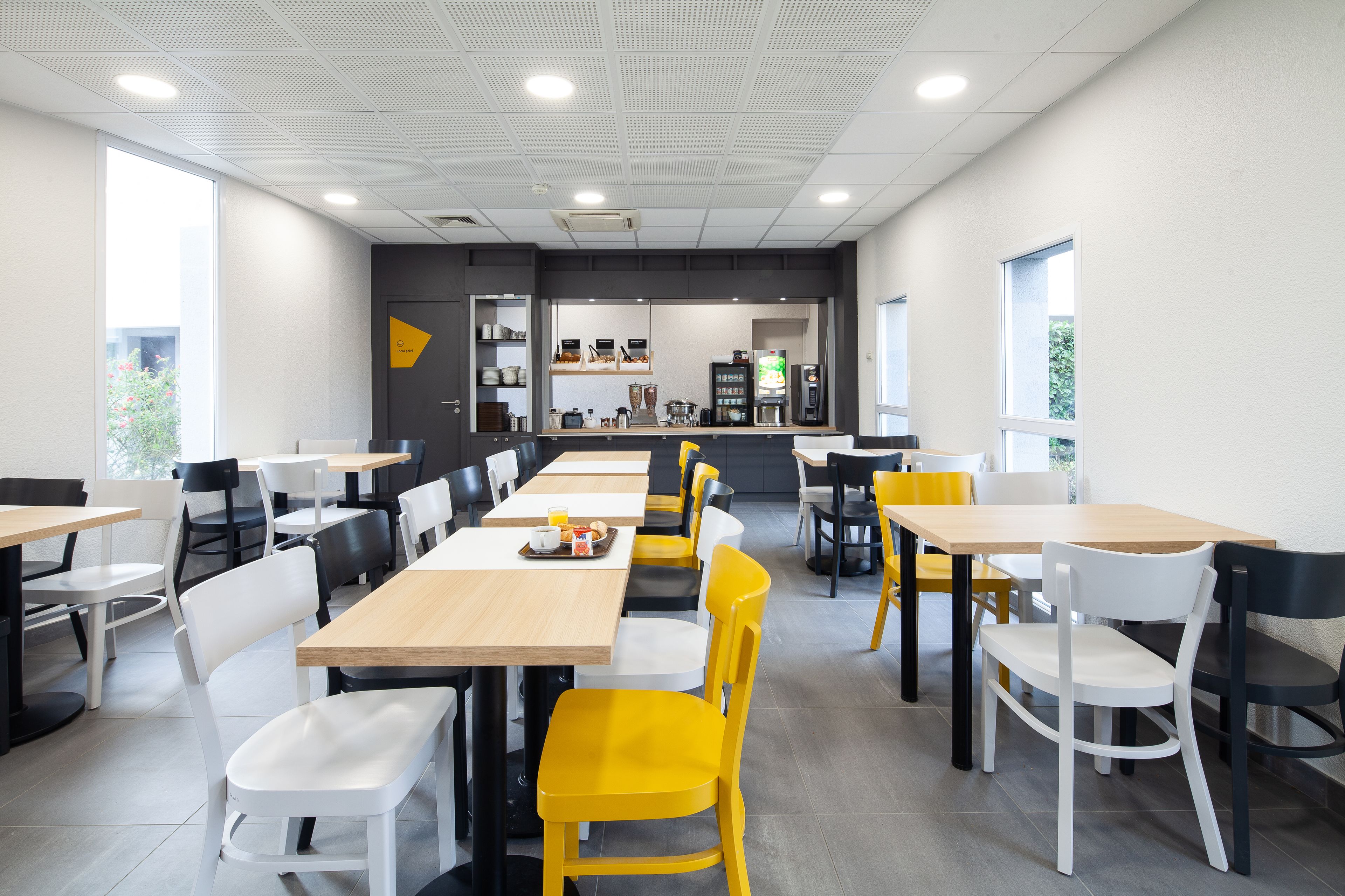A modern dining area with multiple tables and chairs, a kitchen in the background, and large windows allowing natural light.