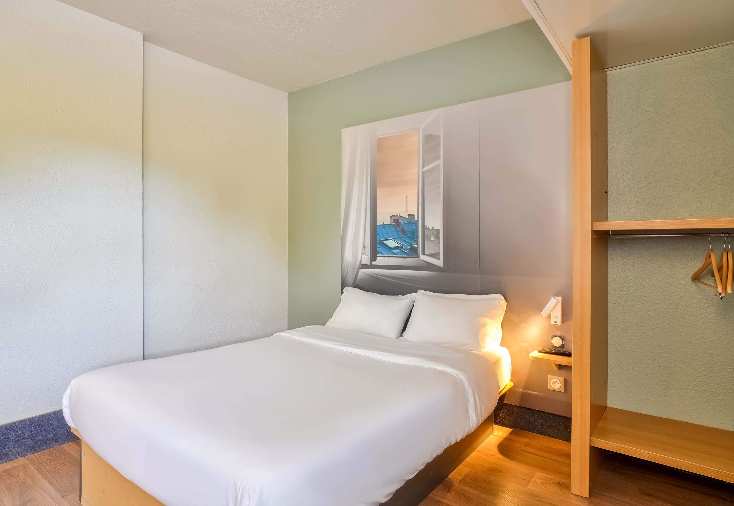 A neatly made bed with white linens, a nightstand with a lamp, and a view of a cityscape through a window.