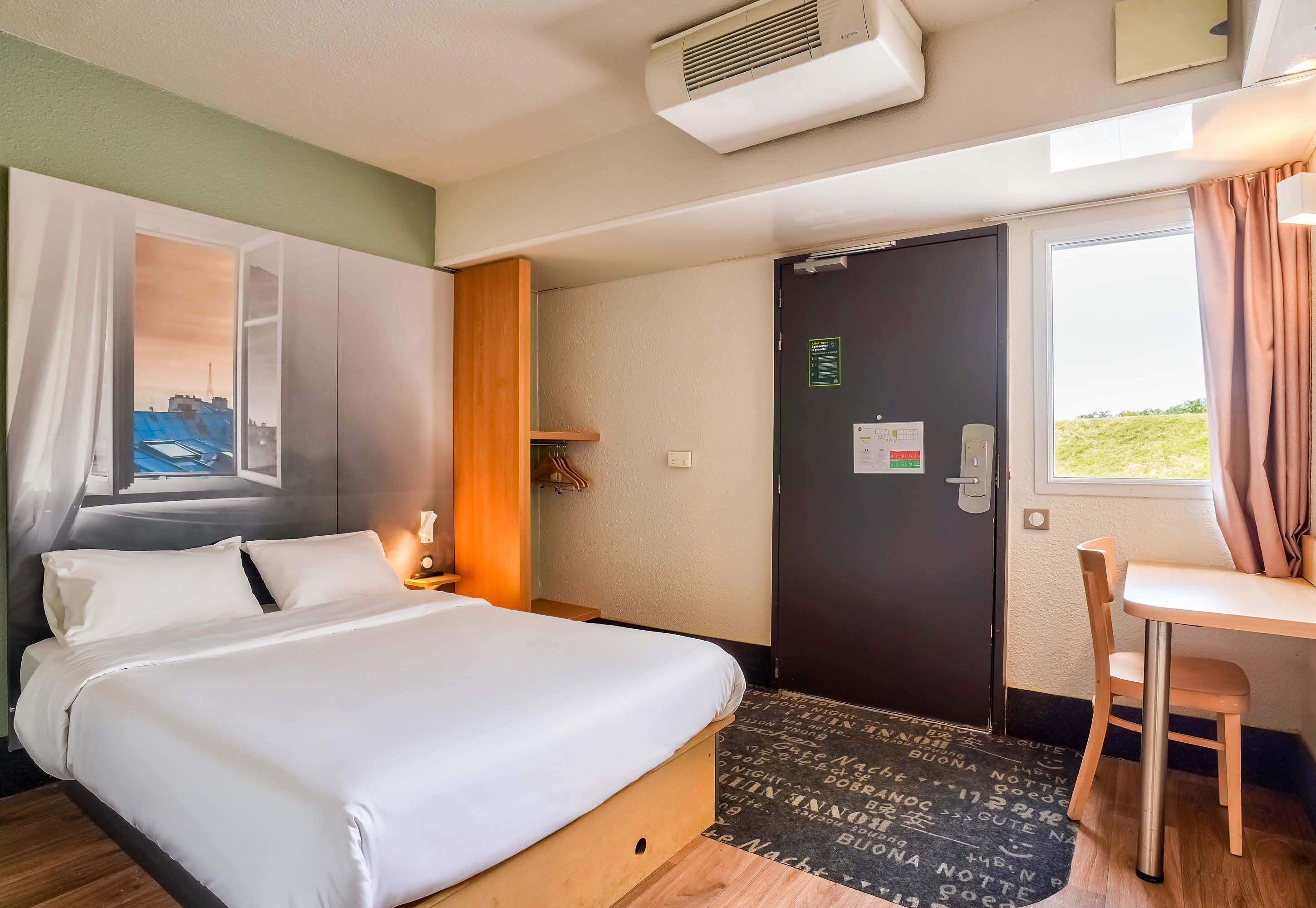 A hotel room with a bed, a desk, a chair, a window with curtains, a door with a sign, and an air conditioning unit.