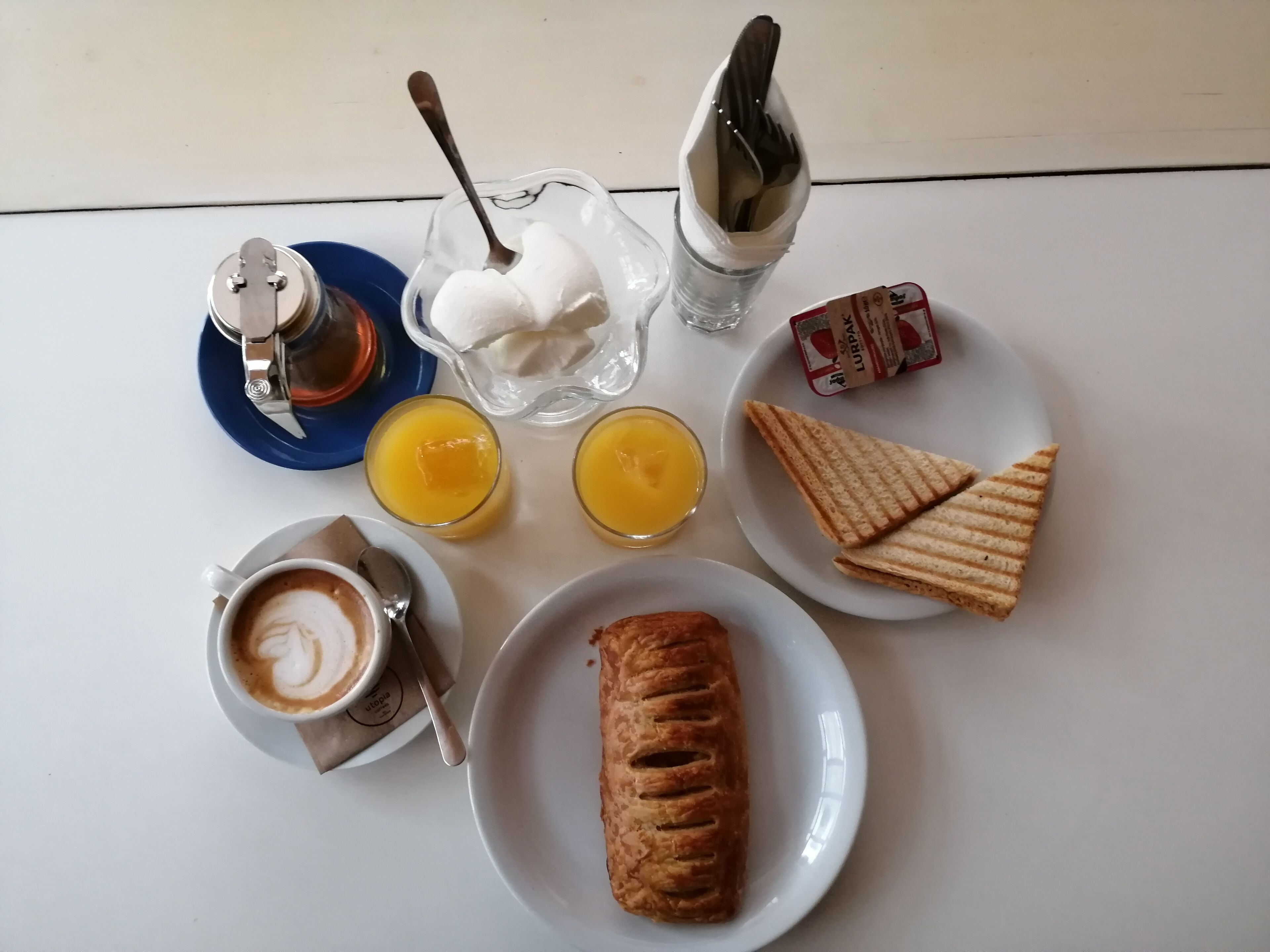 A breakfast setting with a coffee cup, two glasses of orange juice, a sandwich, a pastry, and a sugar container.