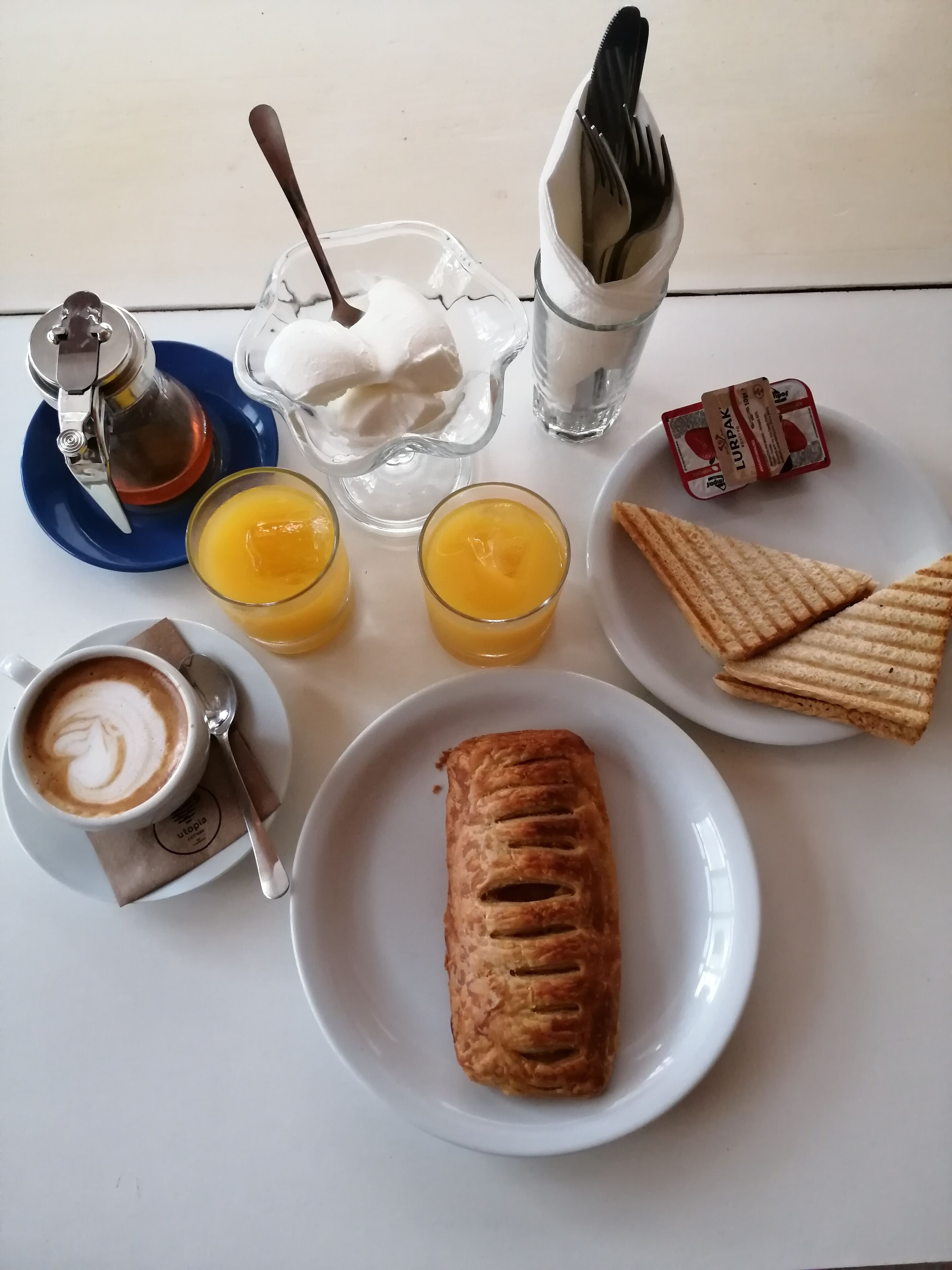 A breakfast setting with a croissant, two glasses of orange juice, a cup of coffee, a small bowl of whipped cream, a packet of butter, a glass of water, and a thermos.