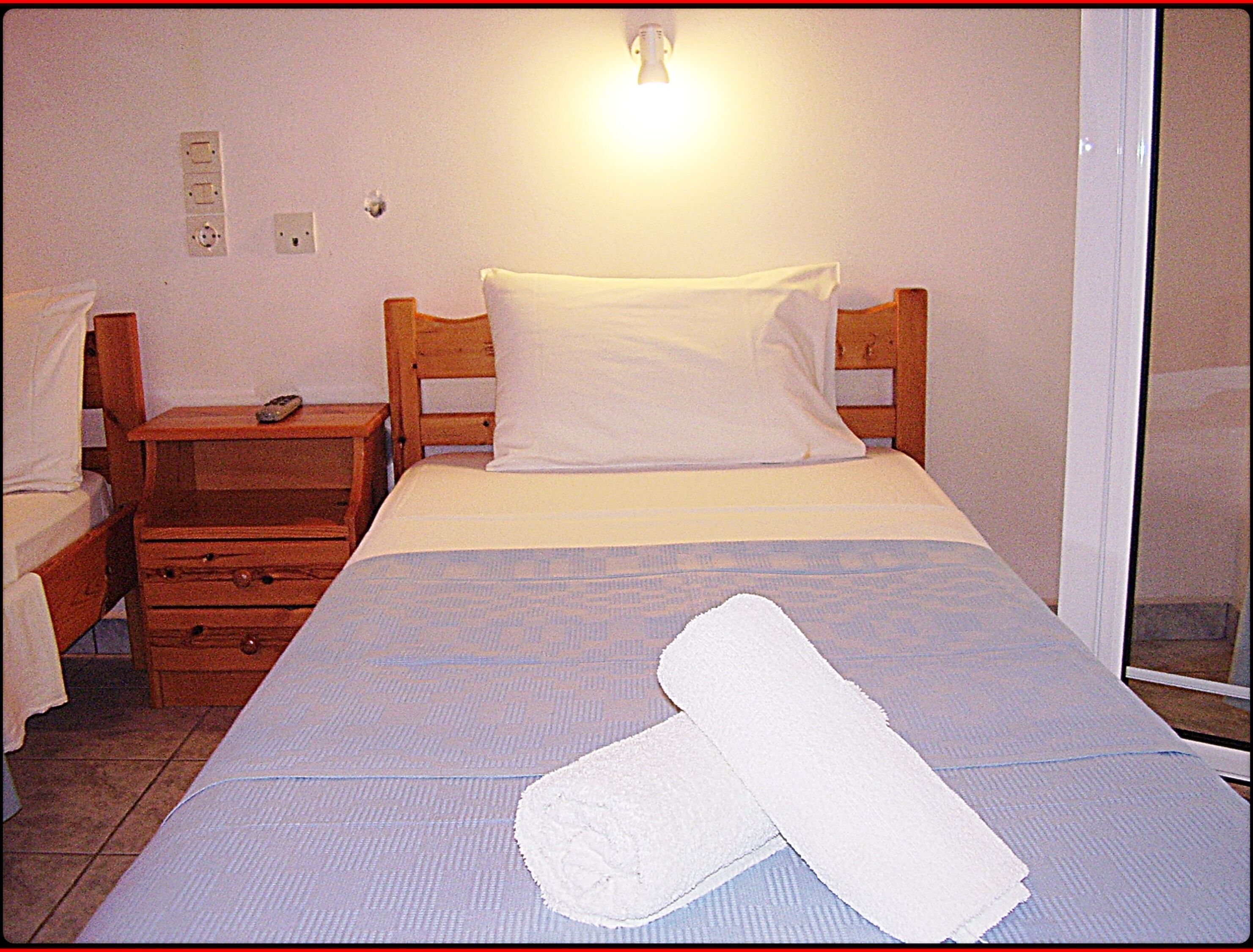 Un letto singolo con cuscino e coperta bianchi, un comodino in legno con un telecomando e una presa elettrica.