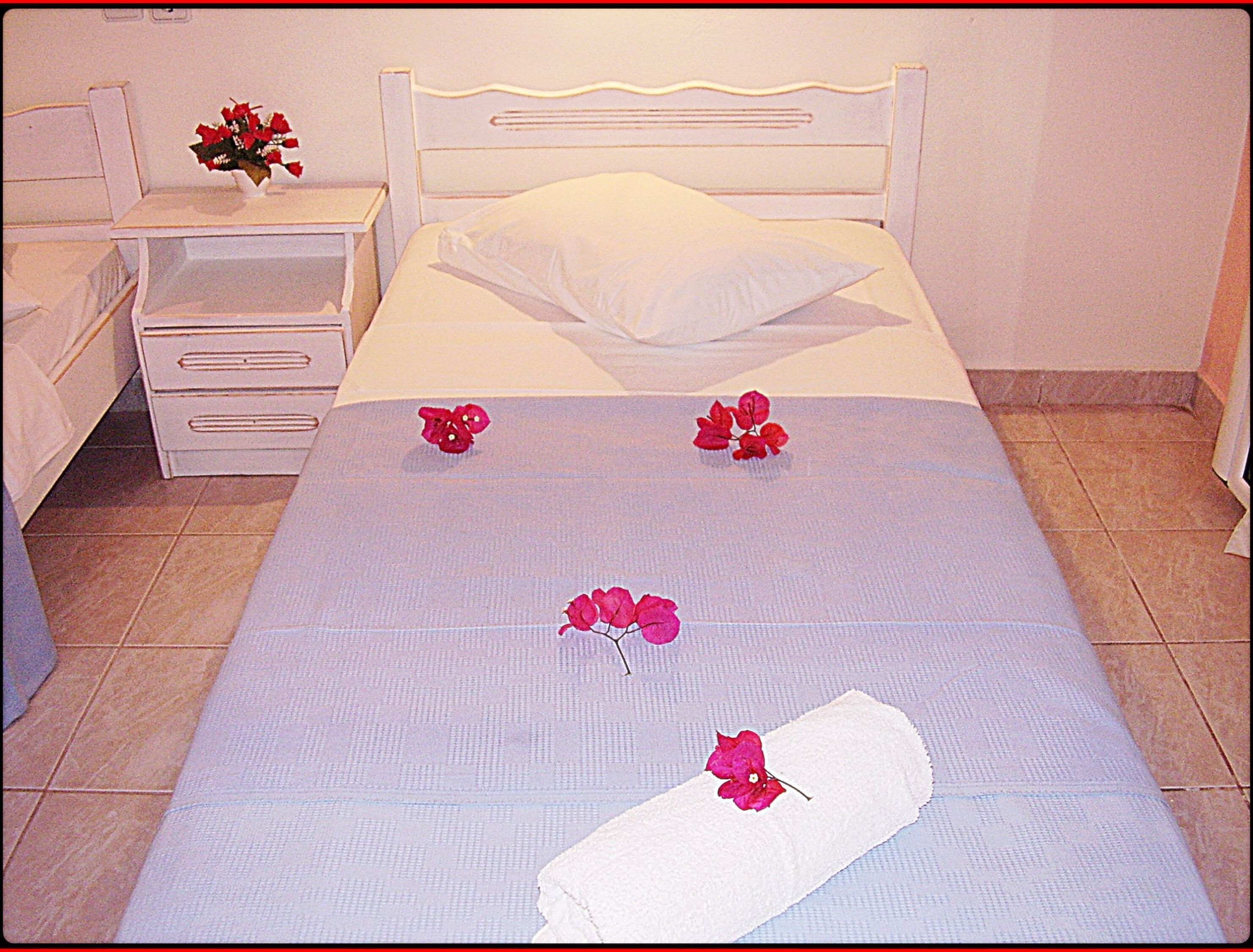A neatly made bed with a white headboard, a white pillow, and a white blanket. There are three red flowers placed on the bed.