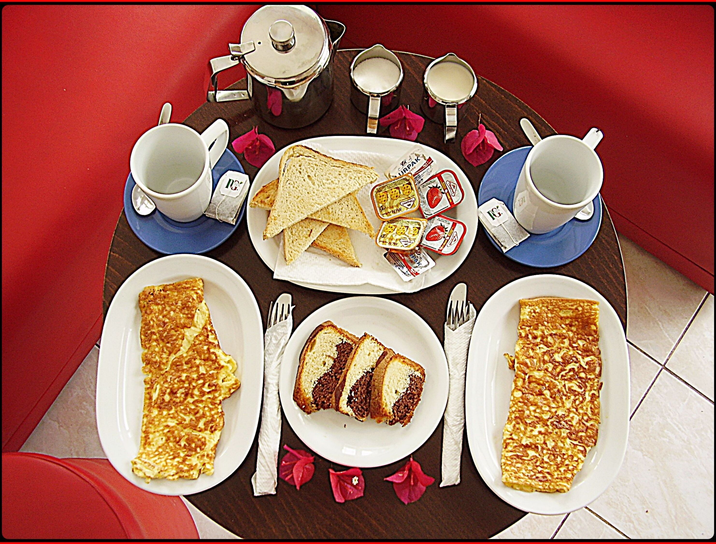 Mesa de café da manhã com diversas opções de alimentos, incluindo torradas, geleia e uma fatia de bolo em pratos, além de xícaras e uma chaleira.