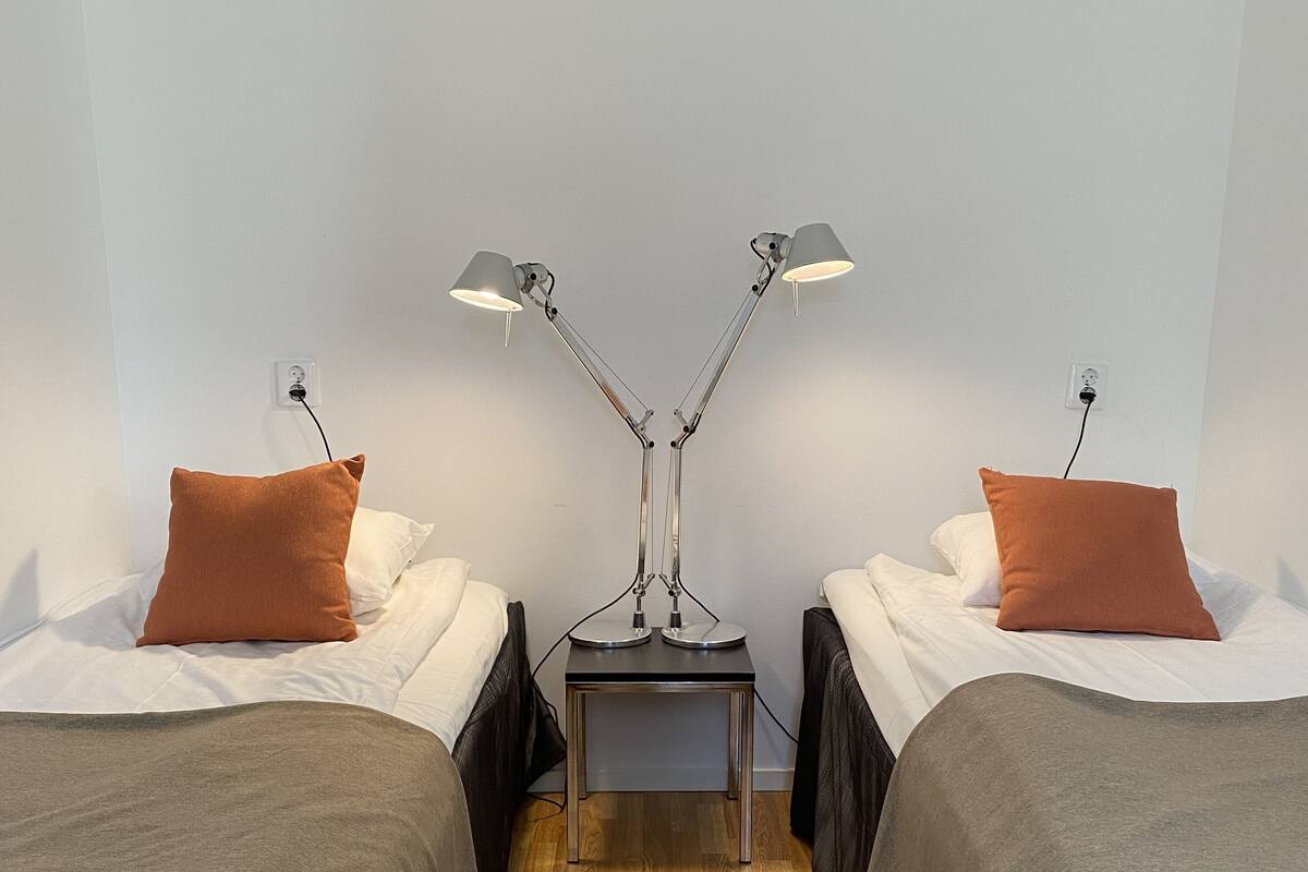 A room with two single beds, each with a brown pillow and white bedding. There is a small table with a lamp between the beds. The floor is wooden, and the walls are white.