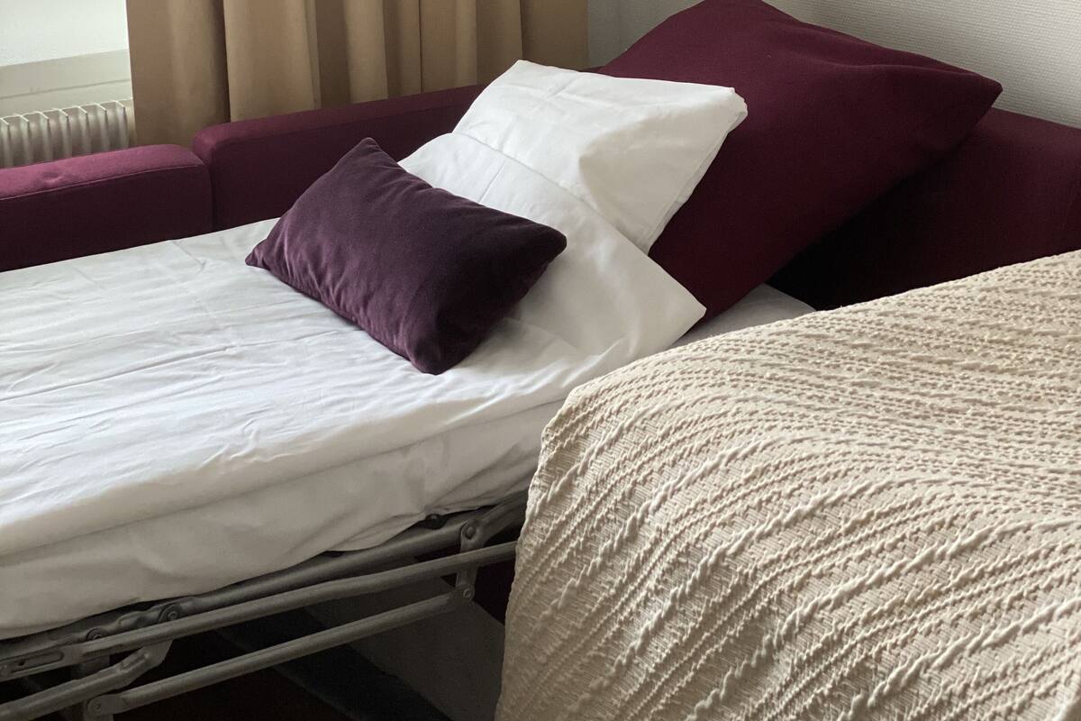 A hotel room with a bed, a window with curtains, and a view of buildings outside.