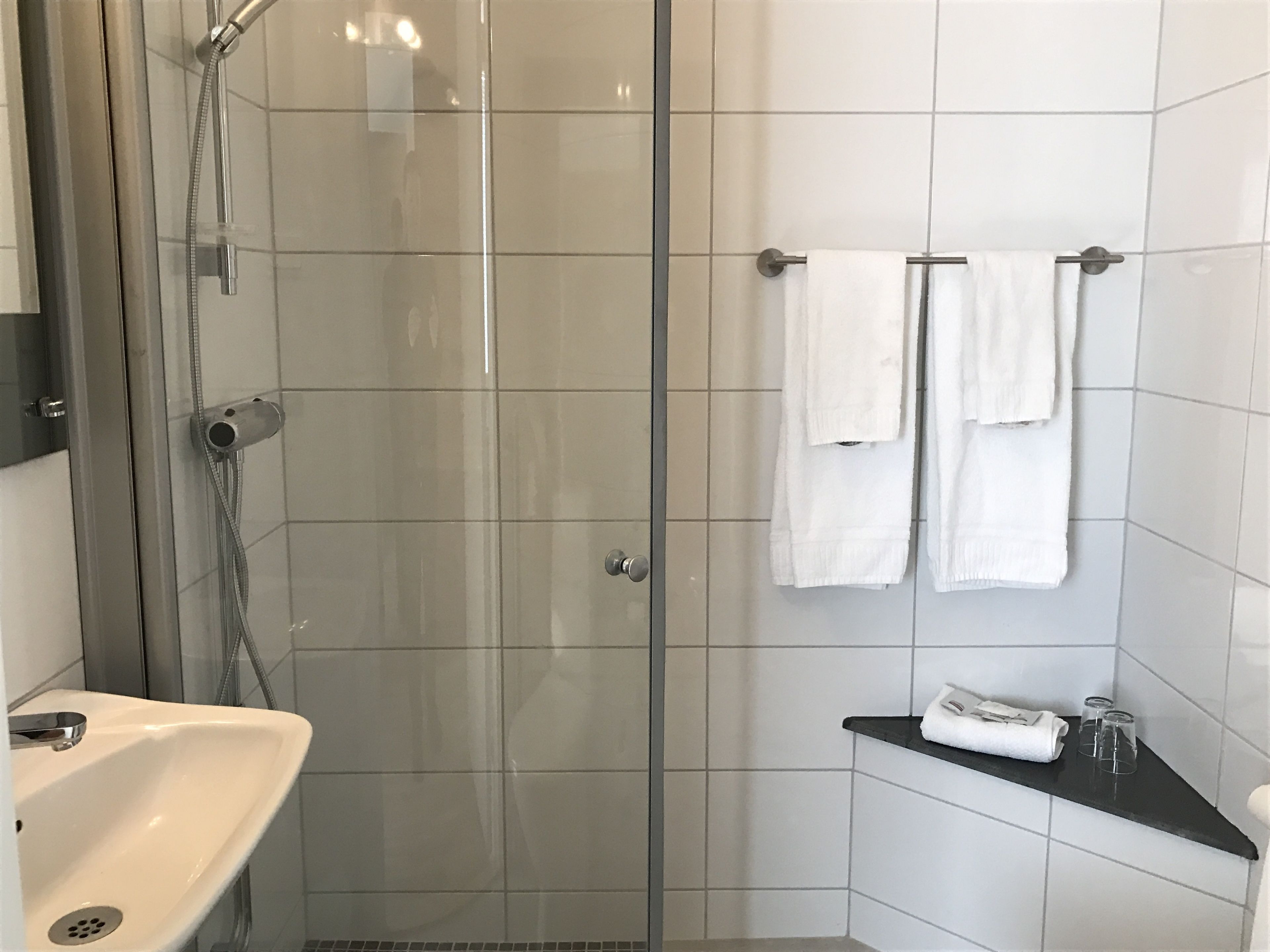 Un baño moderno con ducha envuelta en vidrio, un lavabo blanco y dos toallas blancas en un perchero.