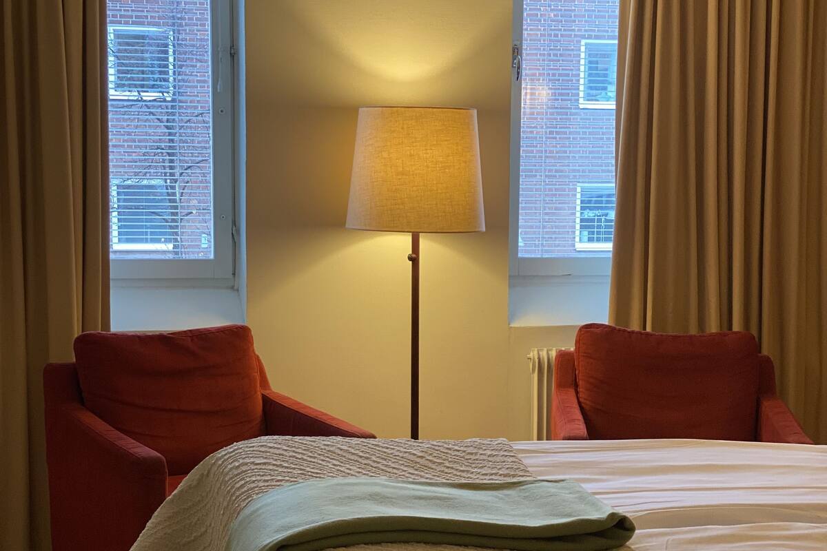 A hotel room with a bed, two red armchairs, a floor lamp, and a window with blinds.