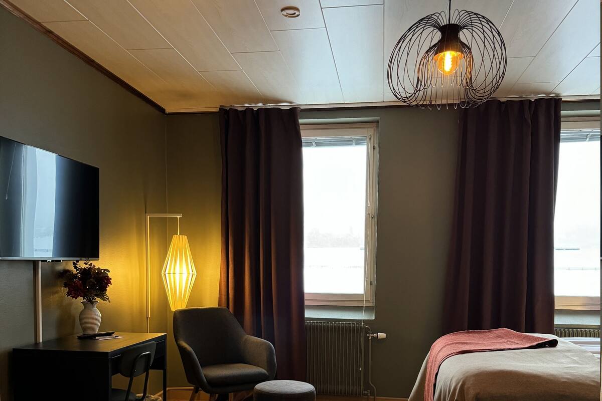 A modern hotel room with a flat-screen TV, a desk with a chair, a round ottoman, a bed with a red blanket, and a unique pendant light.