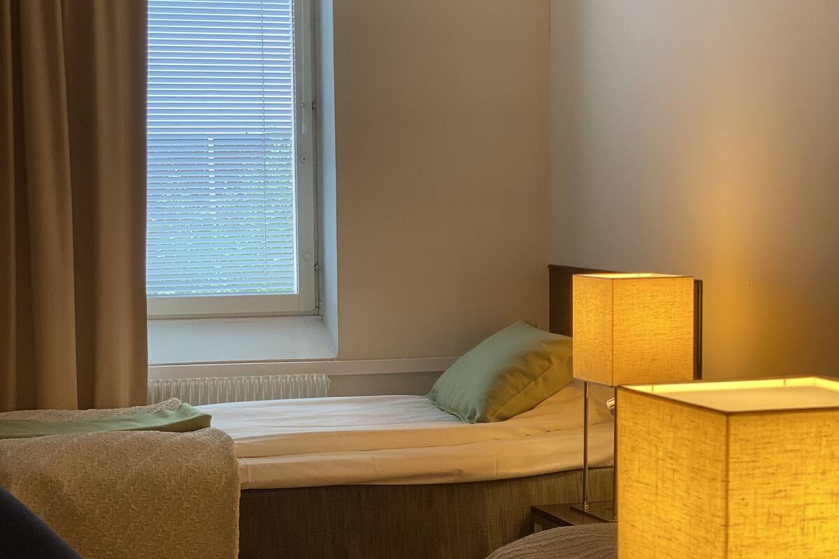 A small, single-room hotel room with a bed, a chair, a table, and a window with blinds.