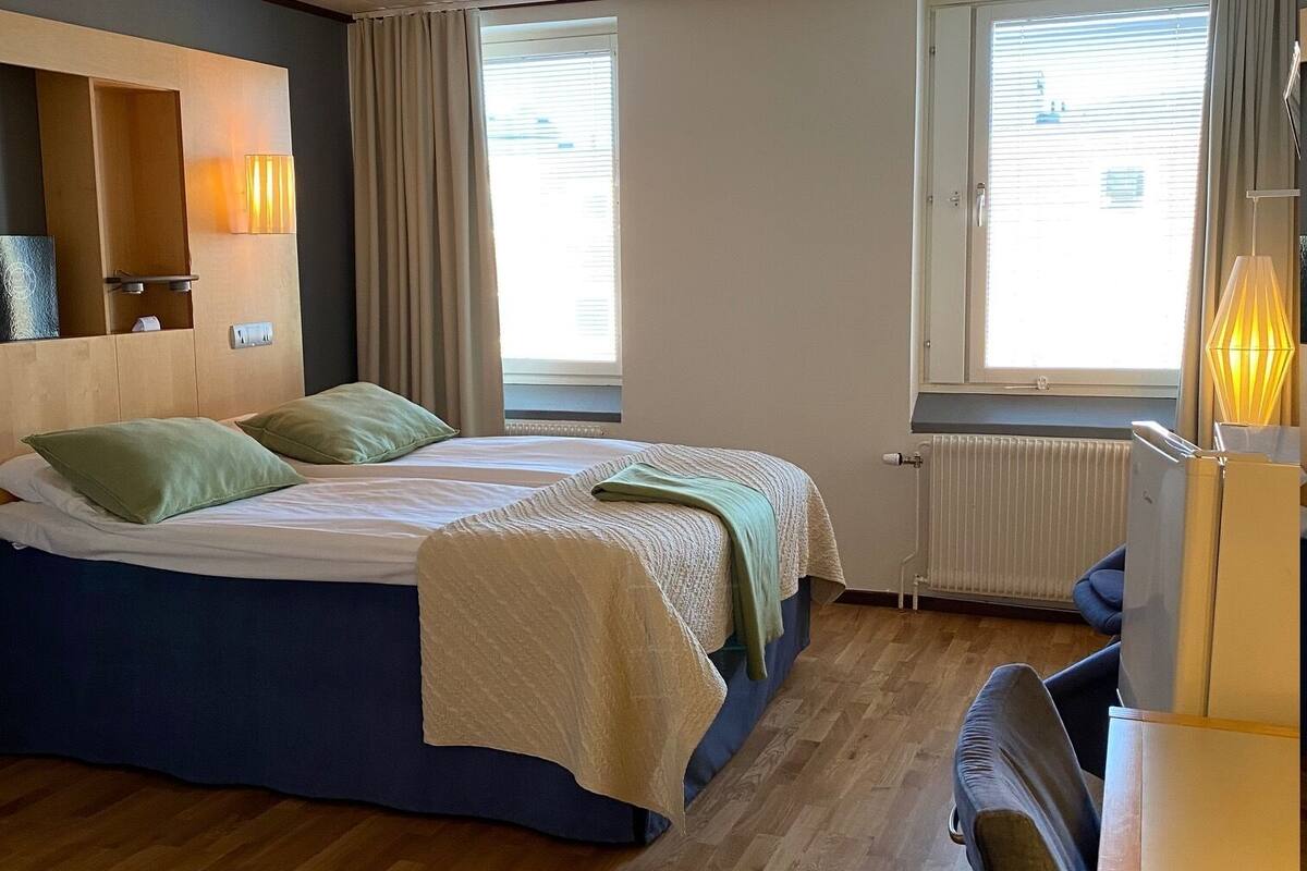 A hotel room with two beds, wooden flooring, a desk, and a window with curtains.