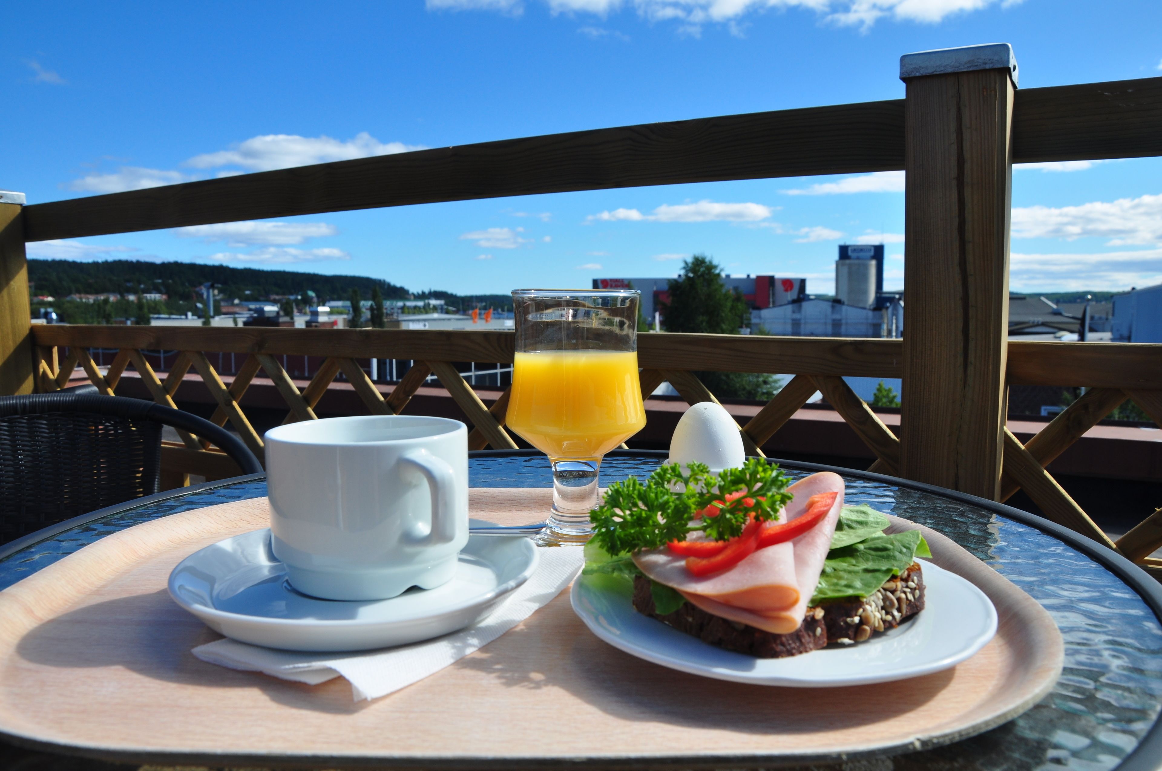 Un plateau de petit-déjeuner avec une tasse, un jus de fruits et un sandwich, posé sur une table d’où l’on peut admirer des bâtiments et des collines.