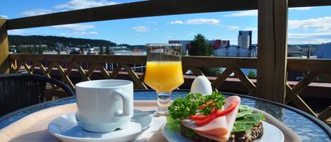 Un plateau de petit-déjeuner avec une tasse, un jus de fruits et un sandwich, posé sur une table d’où l’on peut admirer des bâtiments et des collines.