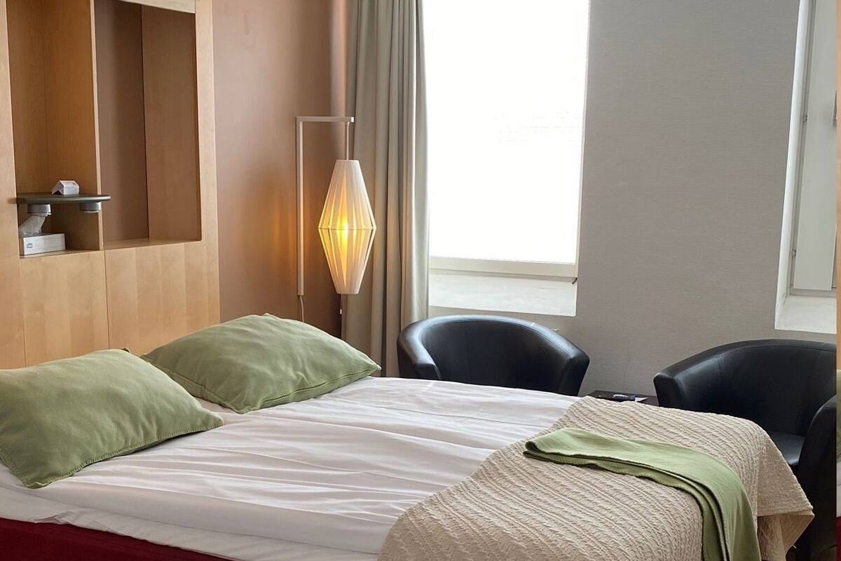 A hotel room with a bed, two chairs, a desk with a lamp, and a window.