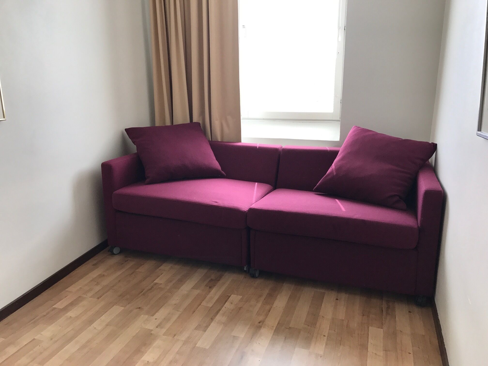 Una habitación de esquina con piso de madera, un sofá púrpura y una ventana con cortinas color beige.