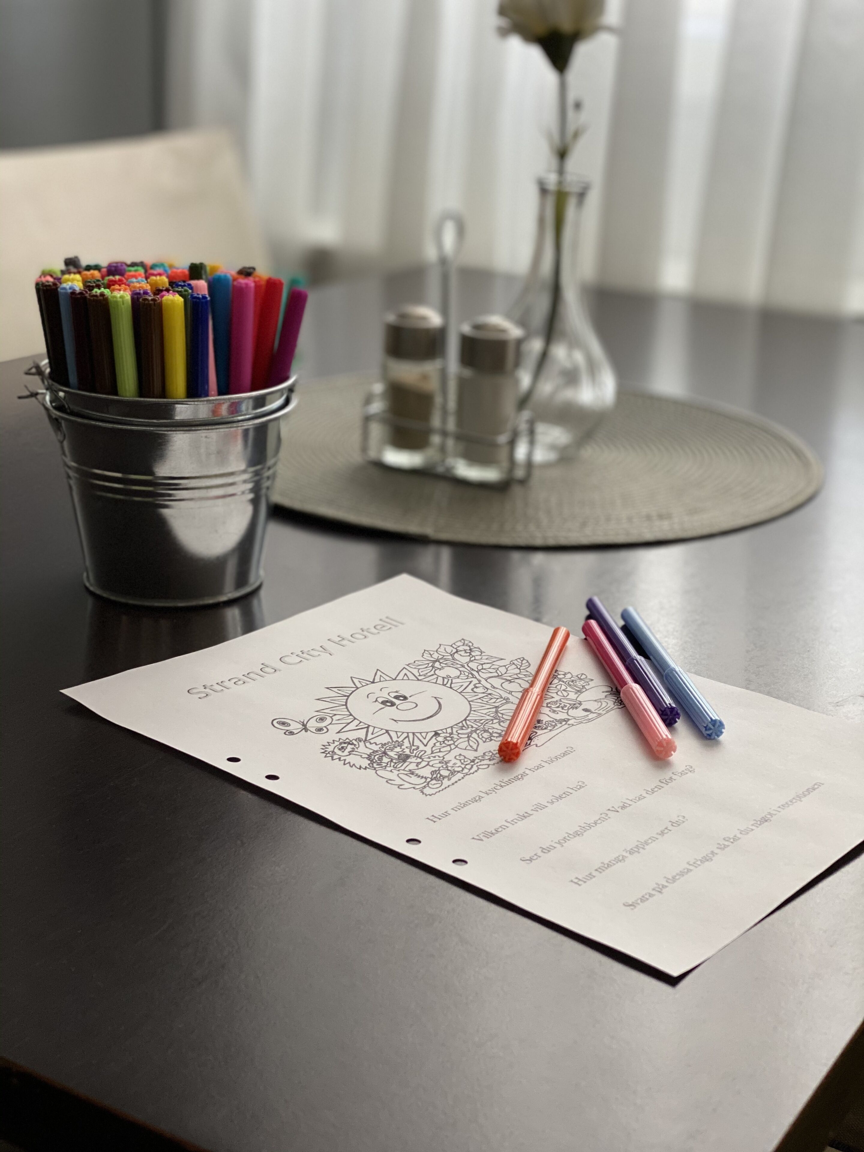 A table with a metal cup containing colorful pens, a glass vase with a single white flower, and a printed sheet with a drawing and text.