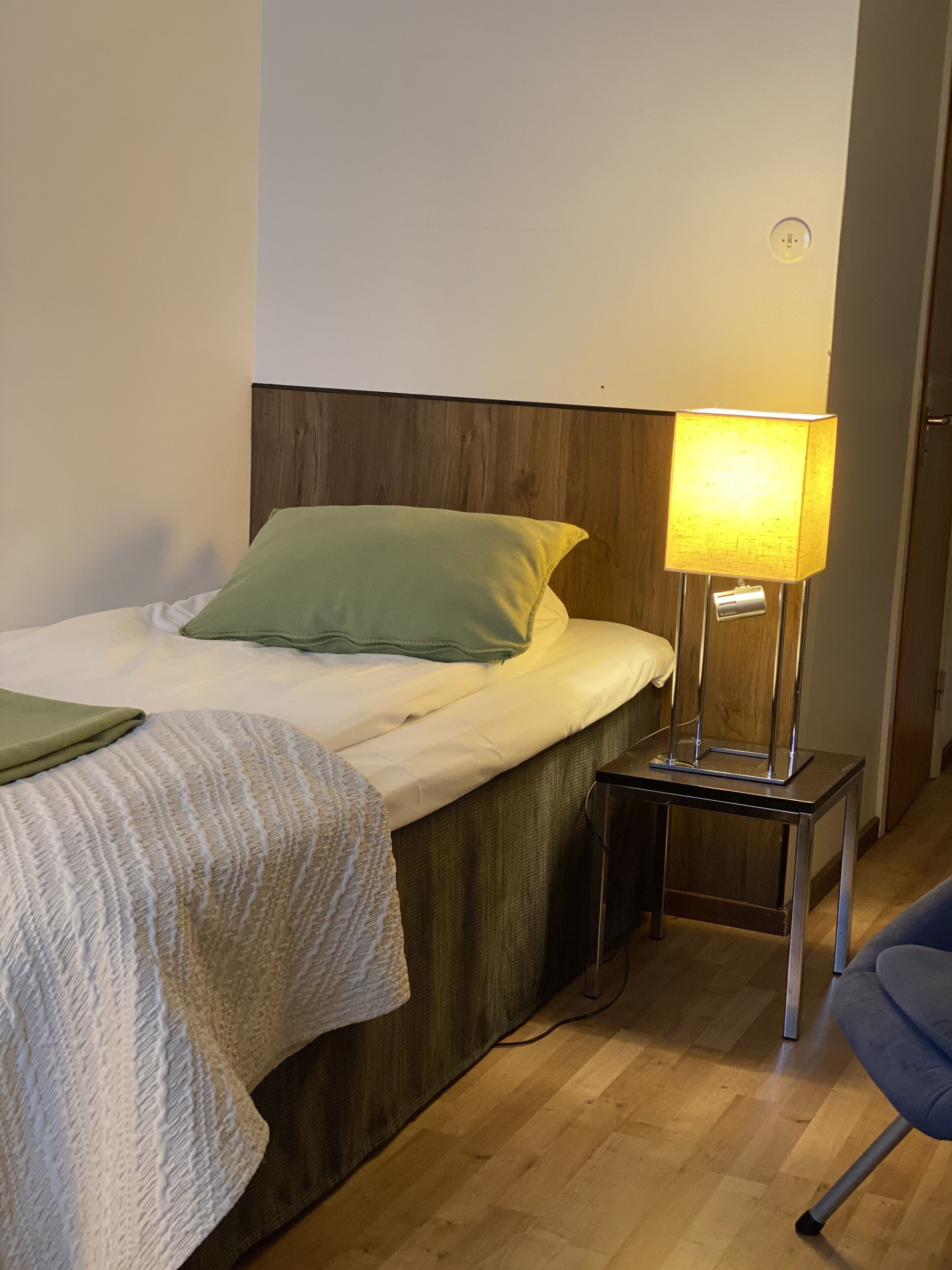 Ein ordentlich bezogenes Bett mit einem grünen Kissen und einer weißen Decke, ein Nachttisch mit einer Lampe und ein Stuhl in einem Zimmer mit Holzboden.