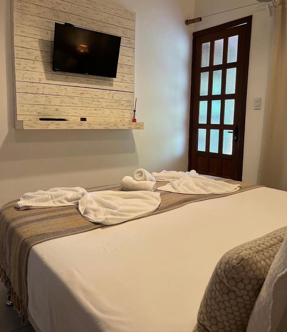 Une chambre d’hôtel avec un lit, un téléviseur fixé sur un mur en panneaux de bois, une porte avec des panneaux de verre dépoli et une serviette sur le lit.