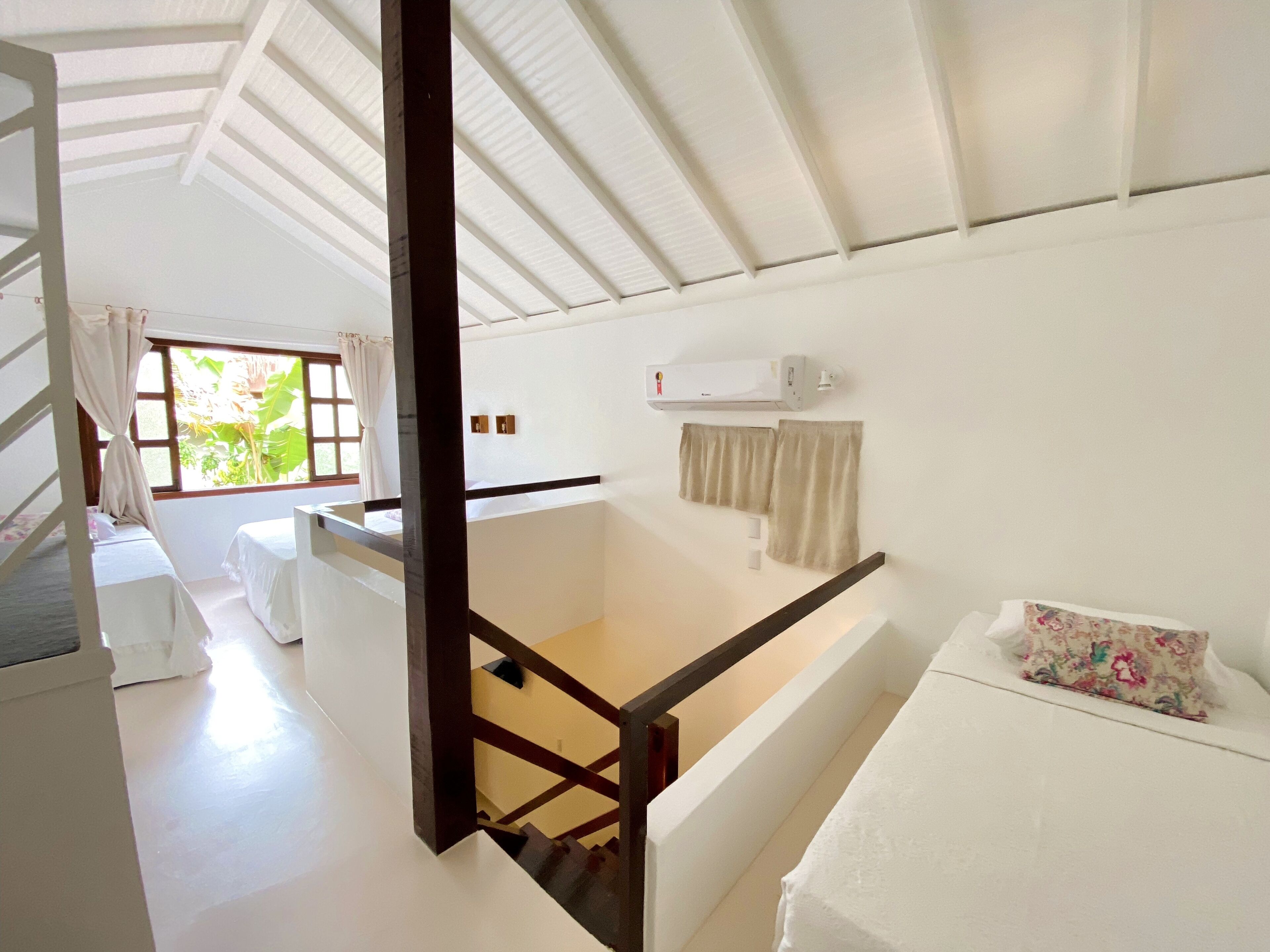 Een ruime kamer met een hoge plafond, een bed met een bloemenkussen, een raam met gordijnen en een witte airconditioning.