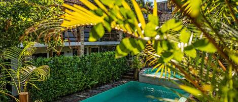 A pool surrounded by lush greenery, wooden deck, and a building with a tiled roof in the background.