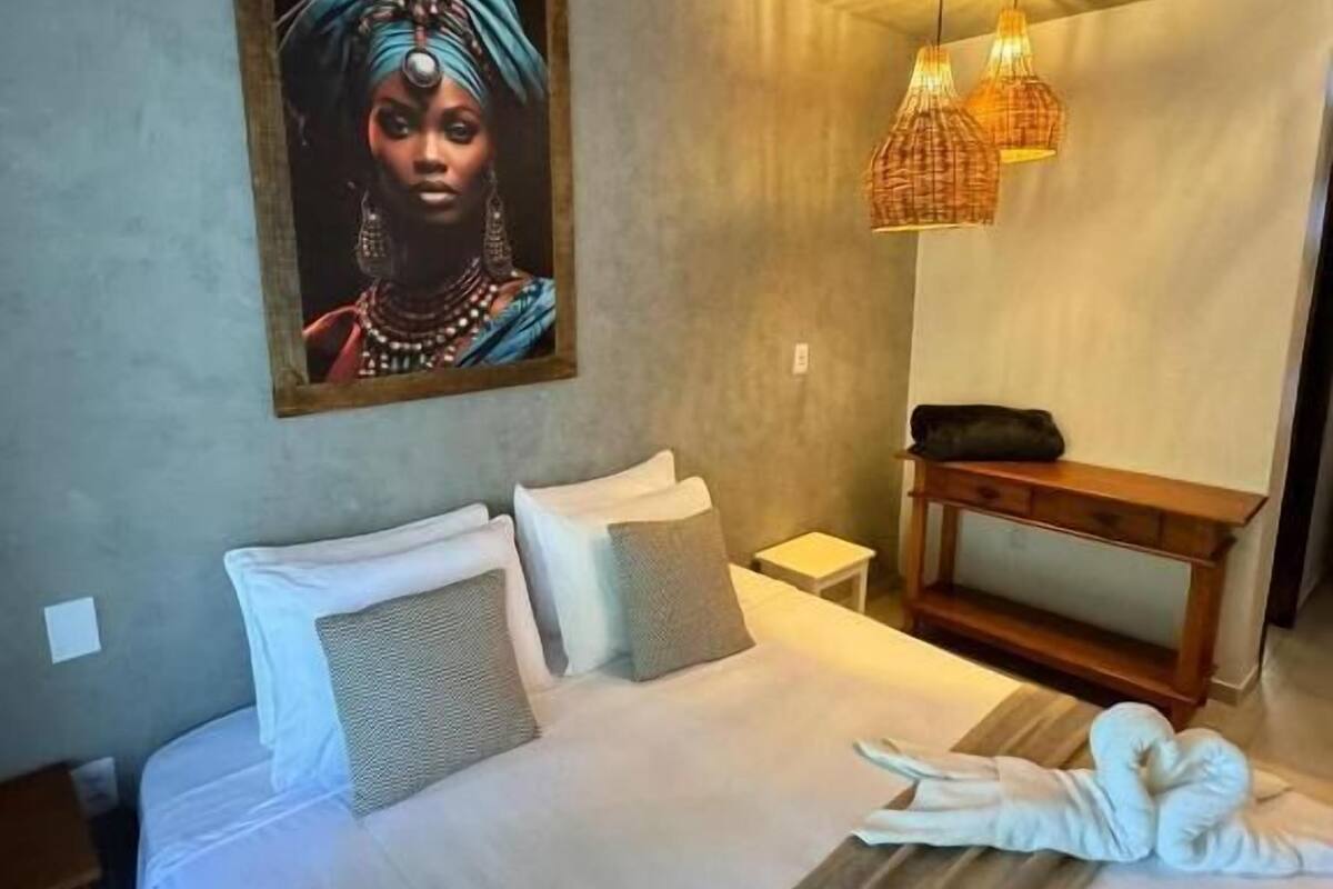 A bedroom with a bed, pillows, a bedside table, a nightstand, a wall-mounted artwork, a hanging light fixture, and a wooden bench.