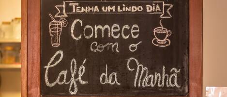 A chalkboard with a message in Portuguese, a coffee machine, and various condiments on a shelf.
