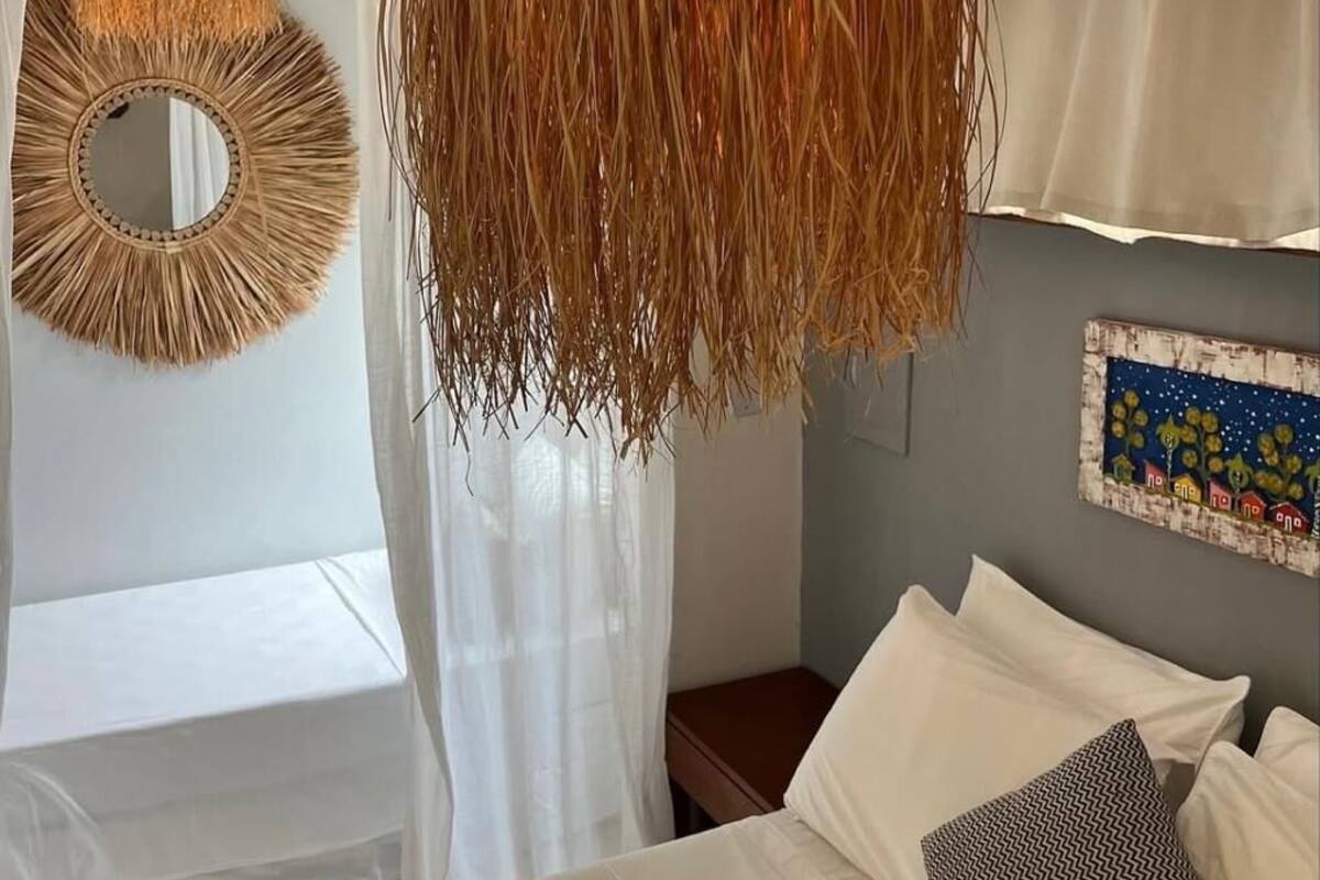A bedroom with a bed, white curtains, a hanging decorative element, a wall mirror, and a wall with a colorful artwork.