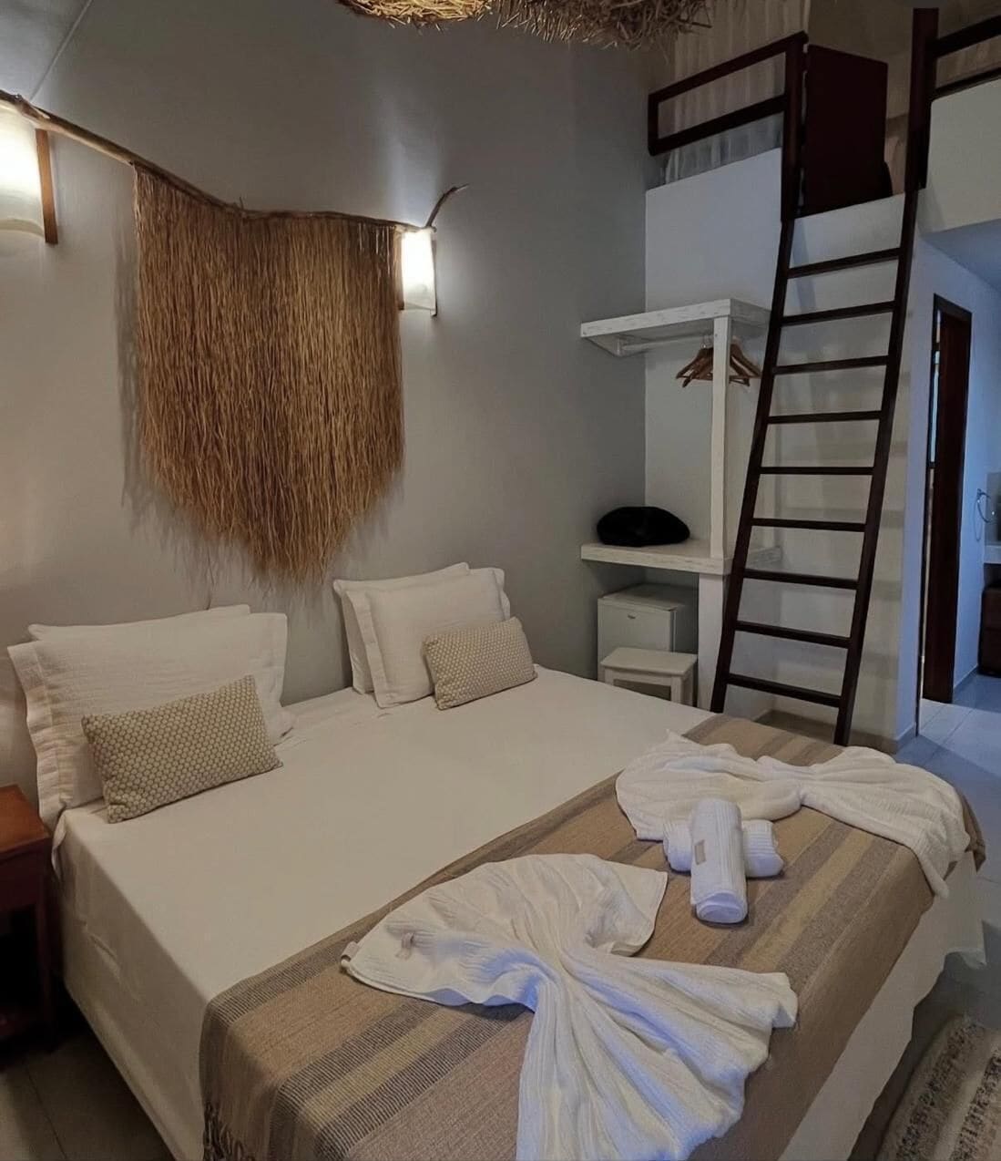 Quarto com cama, almofadas, um adorno pendurado, uma prateleira com um objeto preto e uma escada de madeira.