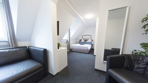 Una habitación de hotel moderna con un sofá de cuero negro, una cama con cabecera blanca, un espejo grande y una planta en maceta.