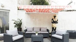 Un área de estar al aire libre con un sofá de ratán gris, dos sillones y una pequeña mesa. Un toldo a rayas ofrece sombra, y hay una planta en maceta a la izquierda.
