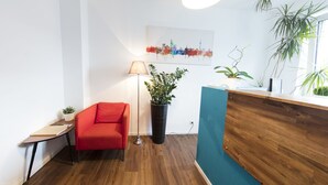 Un área de recepción con un mostrador de madera, una silla roja, una lámpara de pie y un cuadro con una vista de la ciudad.