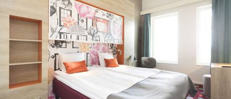 A modern hotel room with a large bed, a headboard with artwork, a desk, and a window with curtains.