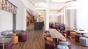 Un hall d’hôtel moderne doté d’un comptoir d’accueil central, de plusieurs espaces de détente avec des tables rondes et carrées, et d’un plancher en bois.
