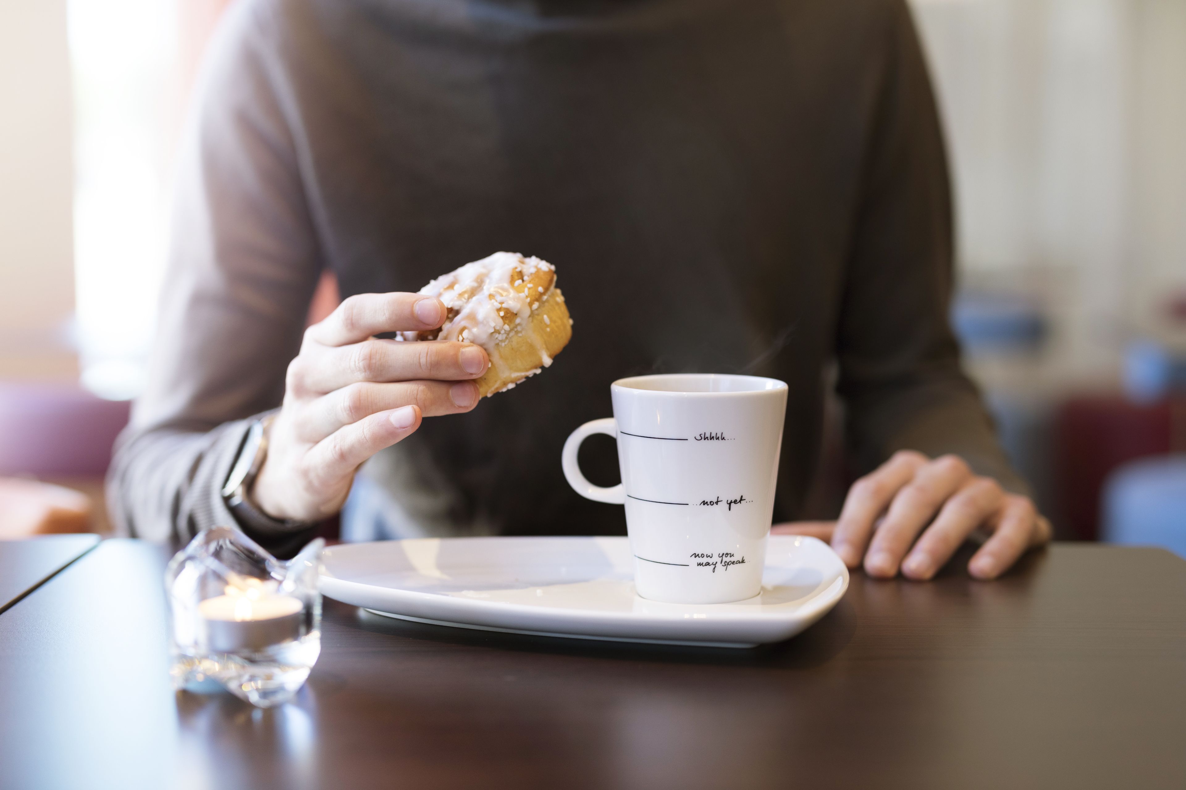 A person holding a donut with icing, sitting at a table with a white mug and a small glass of water.