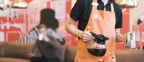A person in an orange apron holding a coffee pot and a metal container, standing in a room with a red wall and a couch.