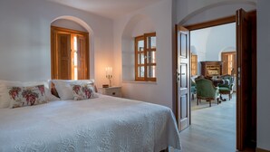 A bedroom with a large bed, a nightstand, a window, and a view of a dining area with chairs. - Casa Santantonio (Santorini)