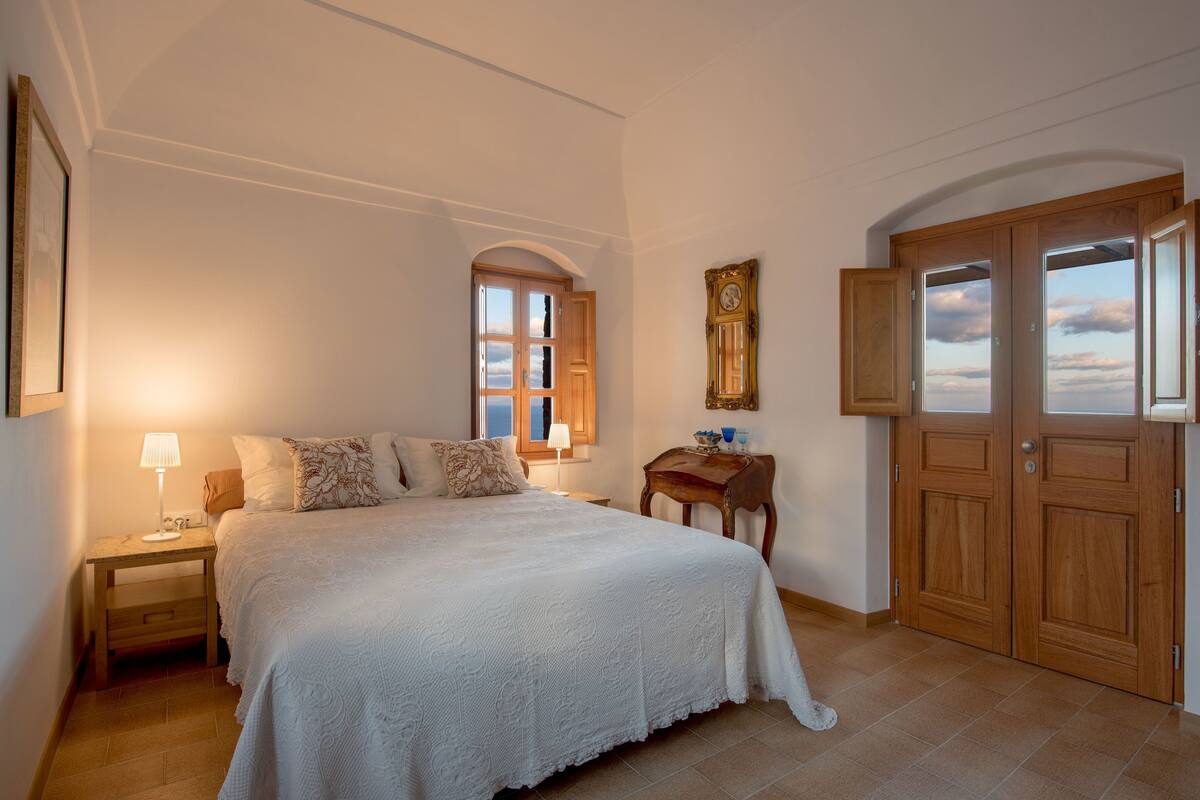 A bedroom with a large bed, a nightstand, a wooden side table, a window with a view, and a door with a mirror.