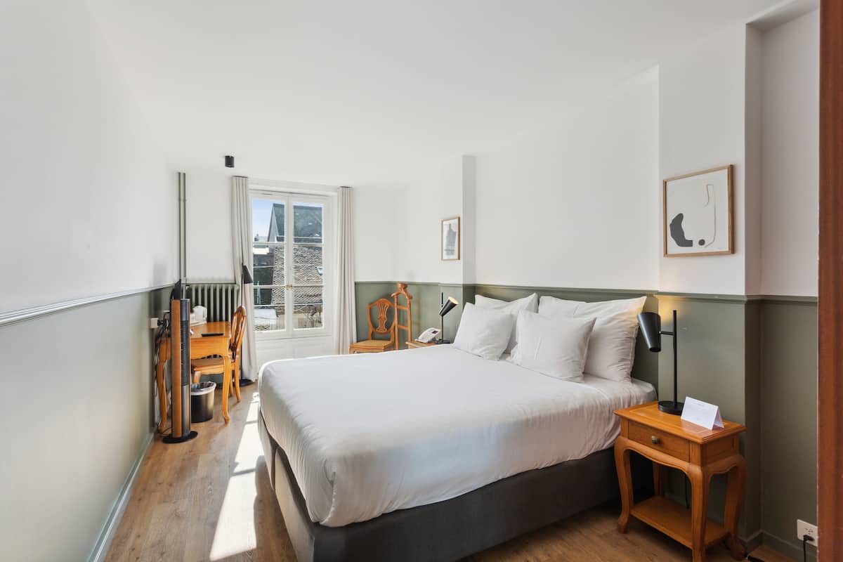 Een slaapkamer met een bed, nachtkastje, stoel en uitzicht op de stad door een raam.