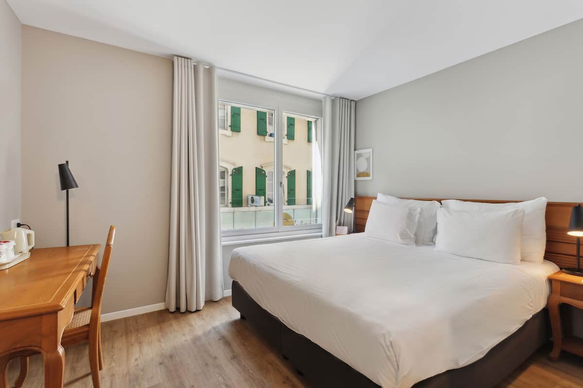 Een hotelkamer met een groot bed, een houten bureau met stoel, een nachtkastje en een raam met gordijnen.