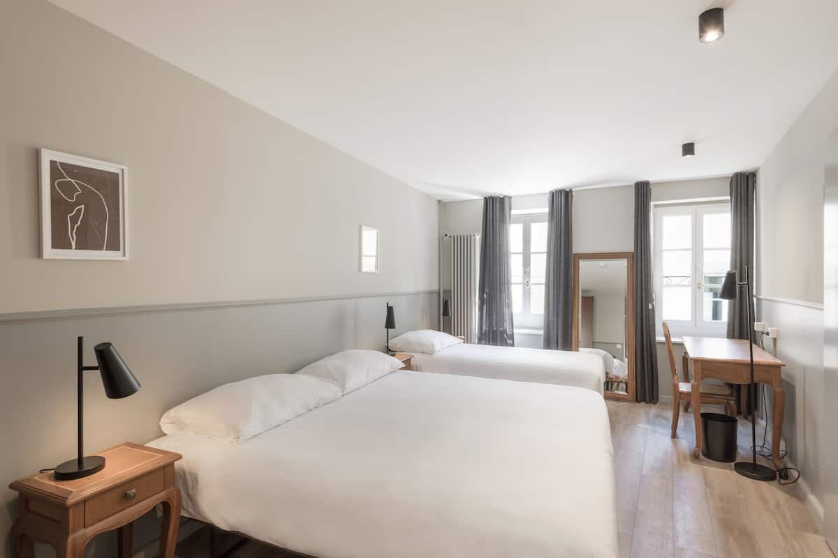 Een hotelkamer met twee bedden, een bureau, een stoel, een lamp, een lijst met een afbeelding aan de muur en een raam met gordijnen.