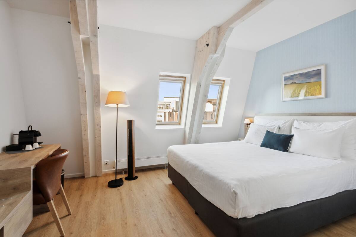 Een moderne hotelkamer met een groot bed, een bureau met stoel, een staande lamp en uitzicht op buiten door twee ramen.