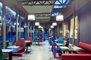 A modern restaurant with glass walls, colorful chairs, and pendant lights.